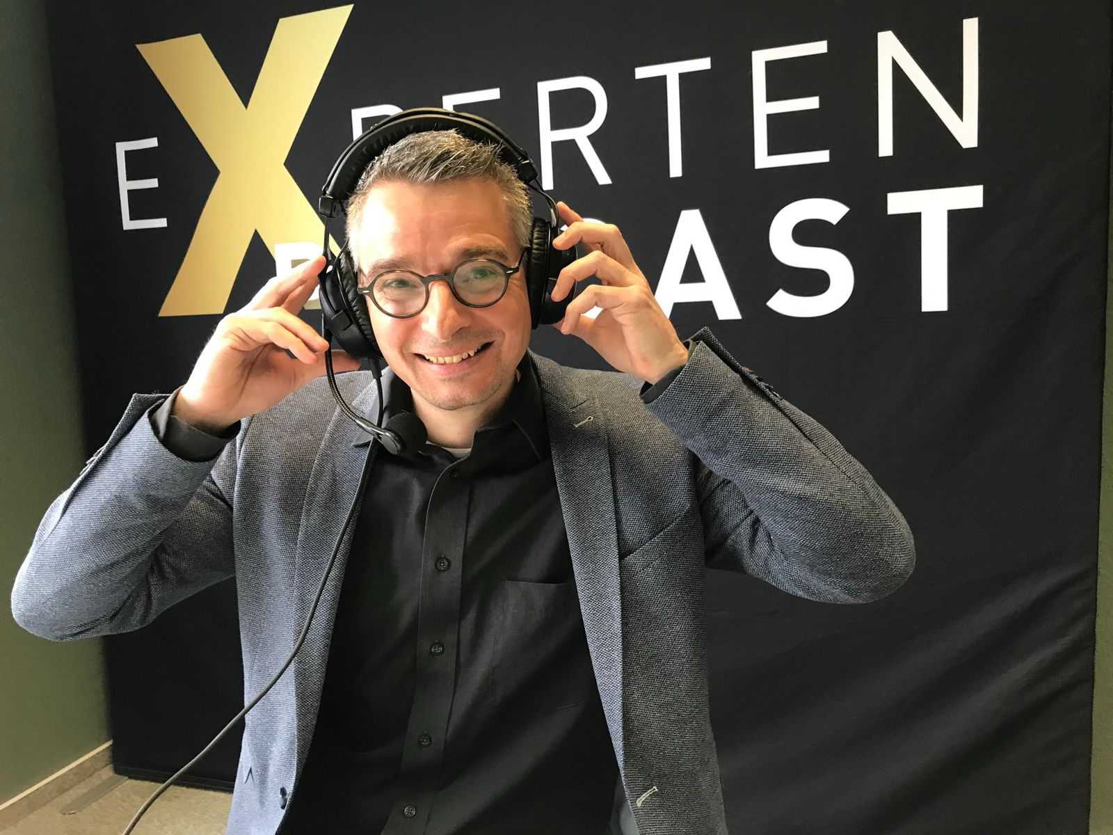 Patrick Schnelle lächelt mit aufgesetzten Kopfhörern in die Kamera, im Hintergrund das Banner des Experten-Podcasts